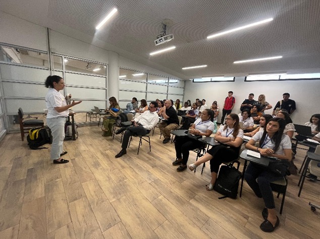 En la imagen se ven unas 20 personas sentadas en pupitres siguiendo la sesión del Modelo APC Colombia, mientras la Coordinadora del Observatorio de Cooperación Internacional expone conceptos técnicos sobre cooperación internacional