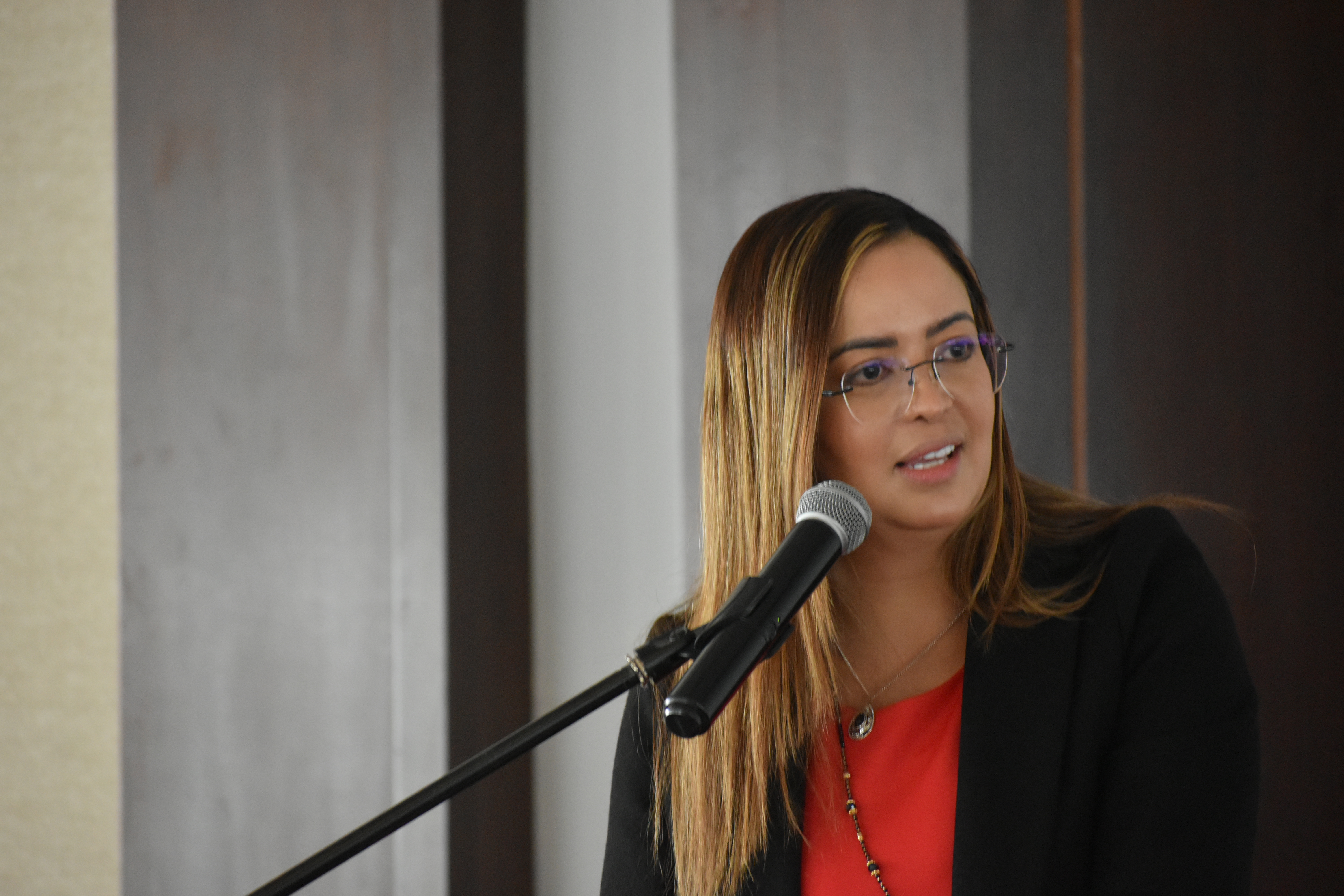 Foto de la directora Viviana Manrique halando por micrófono con una chaqueta color negro y blusa roja