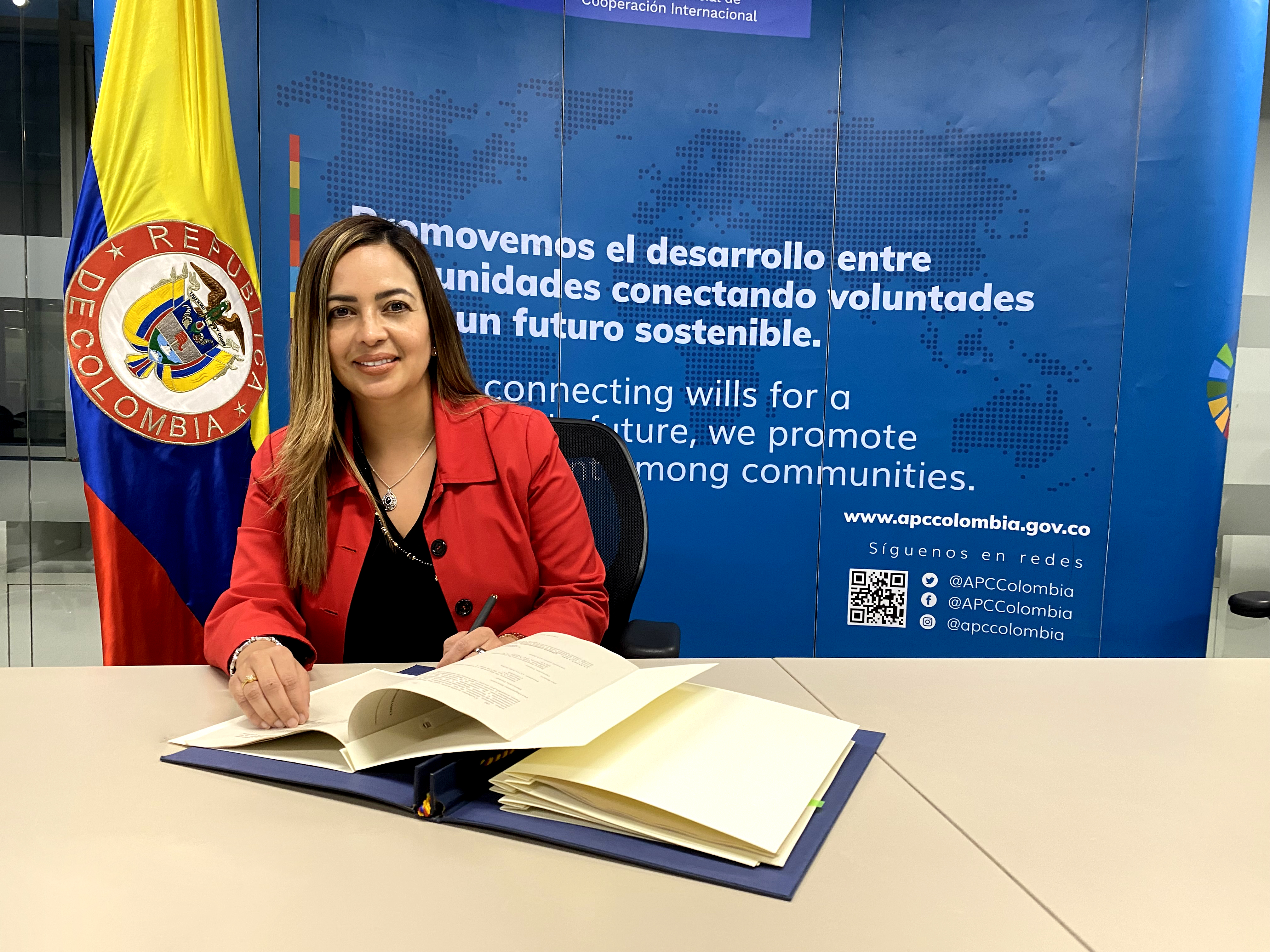 Foto de la directora Viviana Manrique sentada en su escritorio firmando unos documentos con una chaqueta color roja y una blusa negra.