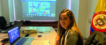 La directora Viviana Manrique Zuluaga en la reunión virtual, frente a ella el computador, al fondo una imagen de los asistentes a la reunión y la bandera de Colombia