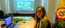 La directora Viviana Manrique Zuluaga en la reunión virtual, frente a ella el computador, al fondo una imagen de los asistentes a la reunión y la bandera de Colombia